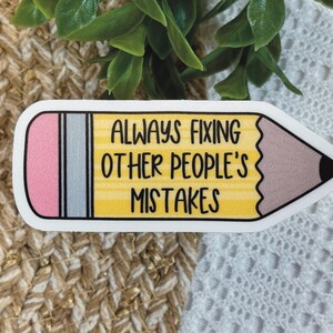 Puede incluir: Una pegatina con forma de l&aacute;piz que incluye una goma rosa, una virola plateada y un cuerpo amarillo con el texto negro ALWAYS FIXING OTHER PEOPLE'S MISTAKES. La punta del l&aacute;piz es marr&oacute;n con una mina negra y tiene un borde blanco.