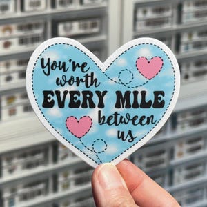 May include: A blue heart-shaped sticker with a white dotted border. The sticker has a blue sky background with white clouds and pink hearts. The text on the sticker reads "You're worth EVERY MILE between us".