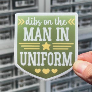 May include: Green and yellow sticker with the text "dibs on the man in uniform" with three yellow hearts at the bottom.