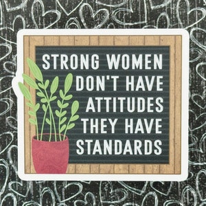May include: A rectangular sticker featuring a black letter board with a light brown wood frame. The text reads: STRONG WOMEN DON'T HAVE ATTITUDES THEY HAVE STANDARDS. A potted plant with light green leaves in a textured red pot sits on the left.