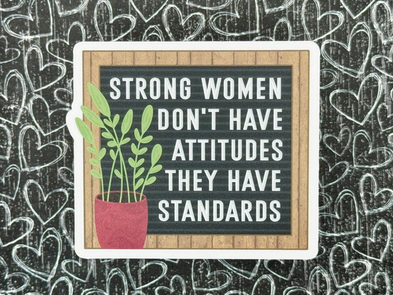 May include: A rectangular sticker featuring a black letter board with a light brown wood frame. The text reads: STRONG WOMEN DON'T HAVE ATTITUDES THEY HAVE STANDARDS. A potted plant with light green leaves in a textured red pot sits on the left.