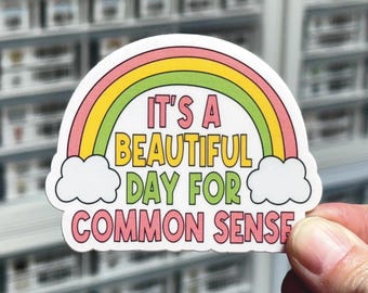 May include: A die-cut sticker featuring a colorful rainbow arching over white clouds. The text "IT'S A BEAUTIFUL DAY FOR COMMON SENSE" is printed in bold, multi-colored letters across the center of the rainbow design against a white background.
