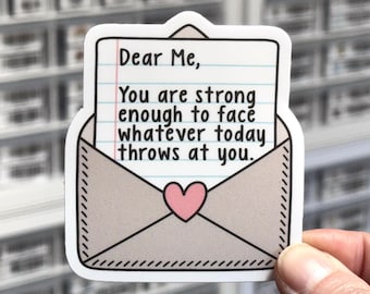 May include: A die-cut sticker featuring a beige envelope with a pink heart seal, containing a piece of lined notebook paper. The paper displays the black text: Dear Me, You are strong enough to face whatever today throws at you.
