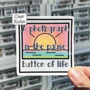 May include: A white sticker with a black border and black text that reads "A photograph is the pause button of life". The sticker has a pink, orange, and yellow sunset graphic with a blue ocean and white waves.