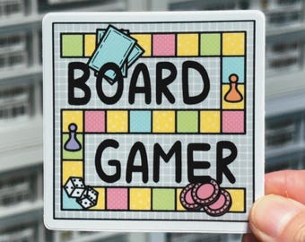 May include: A square sticker featuring a colorful board game path with a grid background. The text BOARD GAMER is printed in large black letters. The design includes illustrations of two playing cards, a game piece, two dice, and three chips.