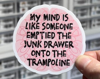 May include: A circular sticker with a pink brain-shaped background featuring a pattern of thin, overlapping lines. The sticker displays the black text: MY MIND IS LIKE SOMEONE EMPTIED THE JUNK DRAWER ONTO THE TRAMPOLINE. The sticker has a white border.