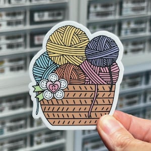 May include: A sticker of a brown wicker basket filled with colorful yarn balls. The yarn balls are blue, yellow, pink, and purple. A white flower with a pink heart in the center is on the side of the basket.