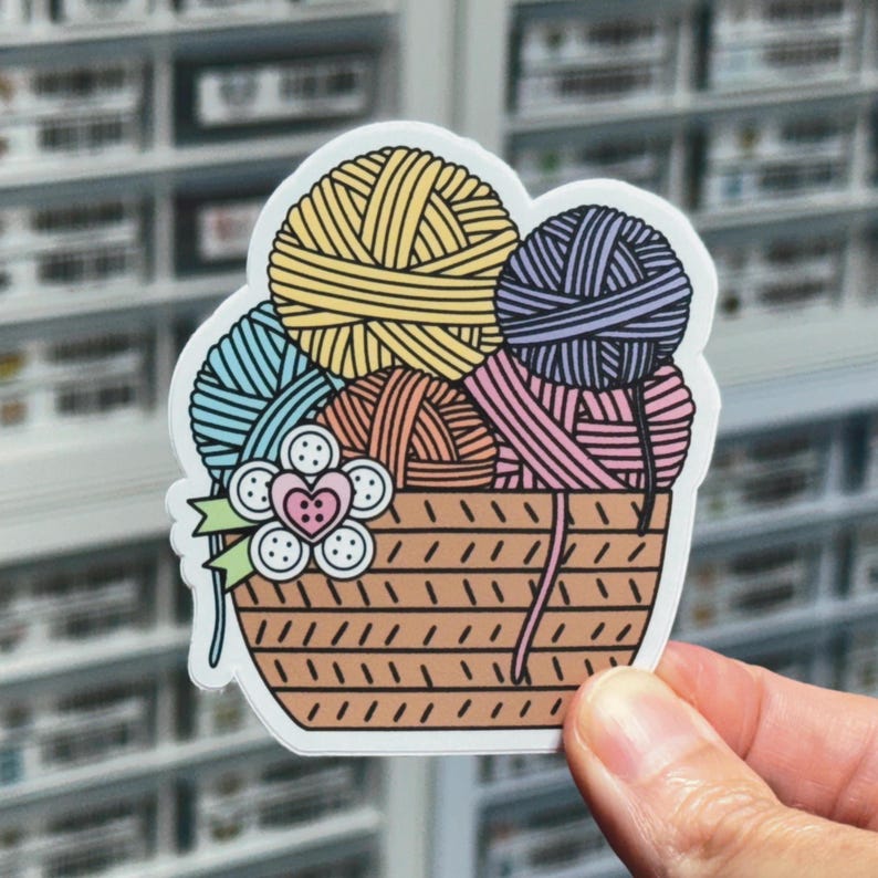 May include: A sticker of a brown wicker basket filled with colorful yarn balls. The yarn balls are blue, yellow, pink, and purple. A white flower with a pink heart in the center is on the side of the basket.