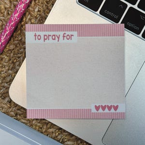 May include: A square sticky note with a light beige center and pink striped borders at the top and bottom. The text "to pray for" is printed in pink on the top border, and four small pink hearts are printed on the bottom border.