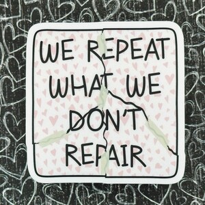 May include: A white sticker with a pink heart pattern and black text that reads "We repeat what we don't repair". The sticker is cracked down the middle.