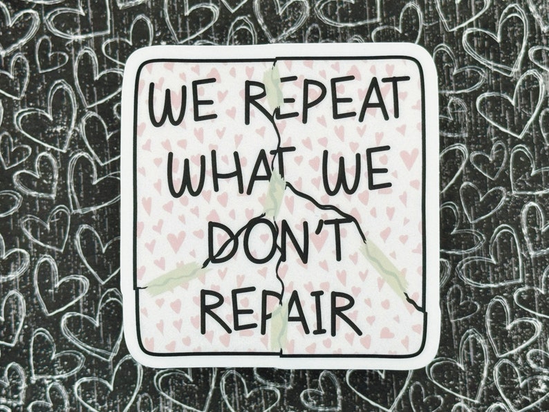 May include: A white sticker with a pink heart pattern and black text that reads "We repeat what we don't repair". The sticker is cracked down the middle.