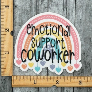 Puede incluir: Una pegatina troquelada con un arco&iacute;ris en tonos pastel, el texto negro "emotional SUPPORT COWORKER" y peque&ntilde;os corazones colgantes. Una regla de madera visible en los bordes indica que la pegatina mide aproximadamente 7,6 cm de ancho.