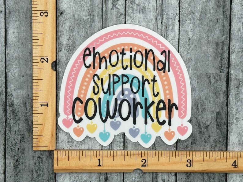 Puede incluir: Una pegatina troquelada con un arco&iacute;ris en tonos pastel, el texto negro "emotional SUPPORT COWORKER" y peque&ntilde;os corazones colgantes. Una regla de madera visible en los bordes indica que la pegatina mide aproximadamente 7,6 cm de ancho.