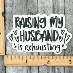 May include: White sticker with black text that reads "Raising my husband is exhausting" with hearts and arrows around the text.