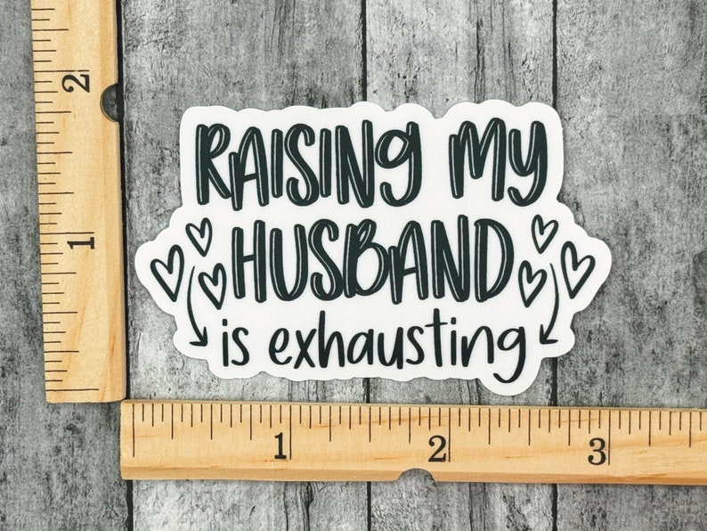 May include: White sticker with black text that reads "Raising my husband is exhausting" with hearts and arrows around the text.