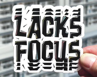 Può includere: Un adesivo in vinile fustellato con il testo LACKS FOCUS in un carattere nero, grassetto e a blocchi. Le lettere hanno un aspetto frammentato e glitch con sottili contorni colorati in blu, giallo e rosa su uno sfondo bianco.