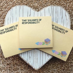 Peut inclure: Trois blocs-notes beiges avec le texte "TINY SQUARES OF RESPONSIBILITY" sont dispos&eacute;s sur une planche en bois blanche en forme de c&oelig;ur. Chaque note a une petite pile de carr&eacute;s color&eacute;s. L'arri&egrave;re-plan est une surface tiss&eacute;e textur&eacute;e.