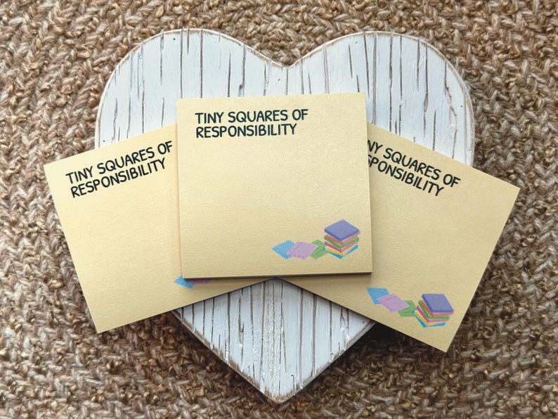 Peut inclure: Trois blocs-notes beiges avec le texte "TINY SQUARES OF RESPONSIBILITY" sont dispos&eacute;s sur une planche en bois blanche en forme de c&oelig;ur. Chaque note a une petite pile de carr&eacute;s color&eacute;s. L'arri&egrave;re-plan est une surface tiss&eacute;e textur&eacute;e.