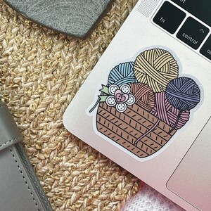 May include: A sticker of a brown wicker basket filled with colorful yarn balls. The yarn balls are in shades of blue, yellow, pink, and purple. There are also some buttons and a flower in the basket.