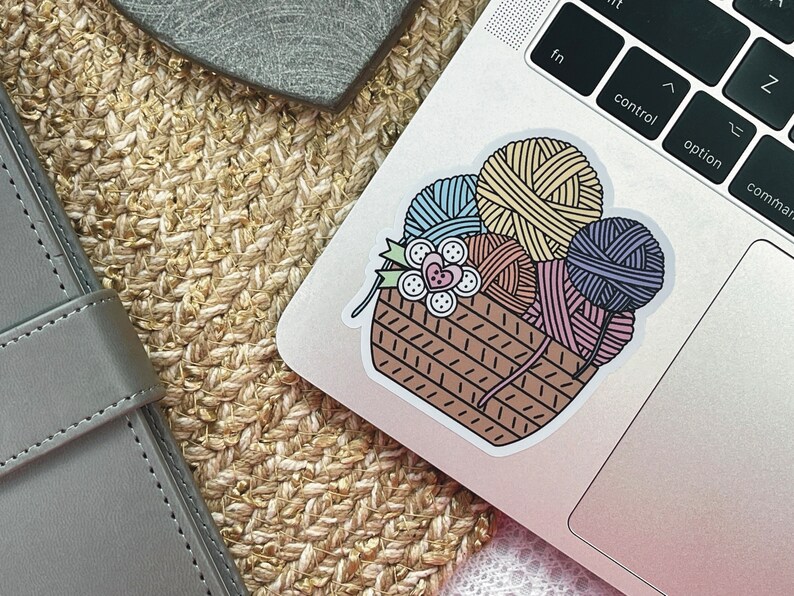 May include: A sticker of a brown wicker basket filled with colorful yarn balls. The yarn balls are in shades of blue, yellow, pink, and purple. There are also some buttons and a flower in the basket.