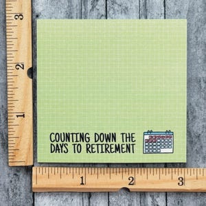 K&ouml;nnte beinhalten: Ein quadratischer, hellgr&uuml;ner Haftnotizzettel mit Gittermuster, bedruckt mit dem schwarzen Text "COUNTING DOWN THE DAYS TO RETIREMENT" und einem kleinen blau-wei&szlig;en Kalendersymbol. Ein Holzlineal zeigt, dass die Notiz etwa 7,6 cm gro&szlig; ist.
