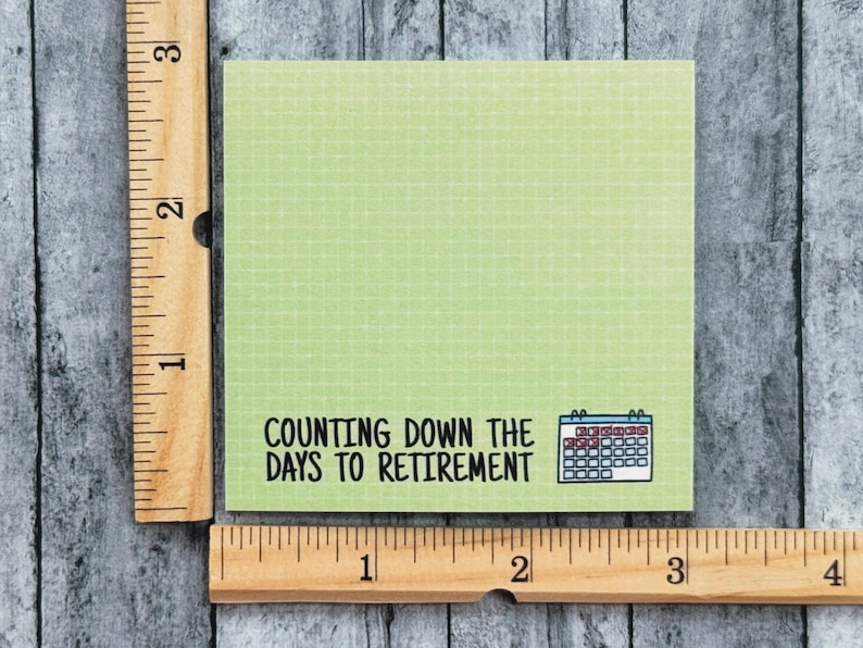K&ouml;nnte beinhalten: Ein quadratischer, hellgr&uuml;ner Haftnotizzettel mit Gittermuster, bedruckt mit dem schwarzen Text "COUNTING DOWN THE DAYS TO RETIREMENT" und einem kleinen blau-wei&szlig;en Kalendersymbol. Ein Holzlineal zeigt, dass die Notiz etwa 7,6 cm gro&szlig; ist.