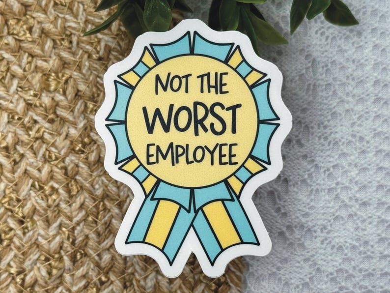 May include: A sticker with a yellow circle and blue ribbons, featuring the text "NOT THE WORST EMPLOYEE" in black. The sticker has a white border and is set against a textured background.