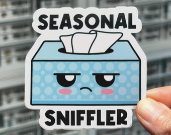 Può includere: Un adesivo fustellato raffigurante una scatola di fazzoletti azzurra con un motivo a pois bianchi e un muso da cartone animato imbronciato con guance rosa. Un fazzoletto bianco sporge dalla parte superiore. Il testo SEASONAL SNIFFLER è in nero.