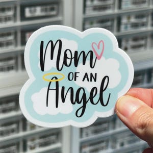May include: A white cloud-shaped sticker with a blue background. The sticker says "Mom of an Angel" with a halo above the word "Angel" and a pink heart next to the word "Mom".