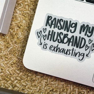 May include: A white laptop sticker with black text that reads "Raising my husband is exhausting" with several small hearts around the text.