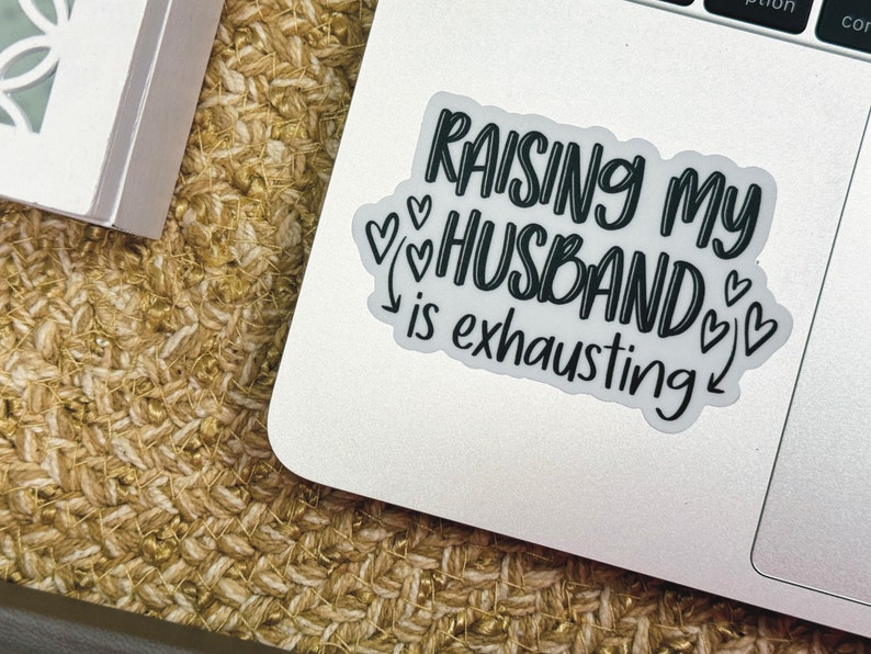 May include: A white laptop sticker with black text that reads "Raising my husband is exhausting" with several small hearts around the text.