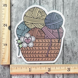 May include: A sticker of a brown wicker basket filled with colorful yarn balls. The yarn balls are in shades of yellow, blue, pink, and purple. There is a small flower with a red heart in the center on the front of the basket.