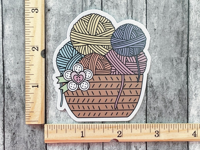 May include: A sticker of a brown wicker basket filled with colorful yarn balls. The yarn balls are in shades of yellow, blue, pink, and purple. There is a small flower with a red heart in the center on the front of the basket.