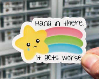 以下が含まれることがあります： 黄色い星と虹が描かれた白いステッカー。星は悲しい顔をしていて、一筋の涙が流れています。ステッカーには「Hang in there. It gets worse.」という文字が書かれています。