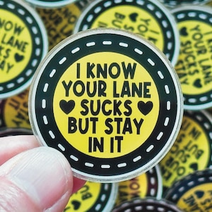 May include: A round, yellow button with the text "I KNOW YOUR LANE SUCKS BUT STAY IN IT" in black. The button has a black border with a road design and white dashed lines. Other buttons are in the background.