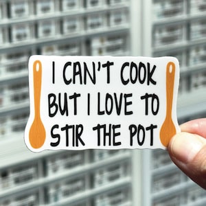 May include: White sticker with the text "I CAN'T COOK BUT I LOVE TO STIR THE POT" in black, with orange wooden spoon illustrations on each side. The sticker is held by a hand.