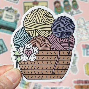 May include: A sticker of a brown basket filled with colorful yarn balls. The basket has a white flower with a pink heart in the center. The yarn balls are blue, yellow, pink, and orange.