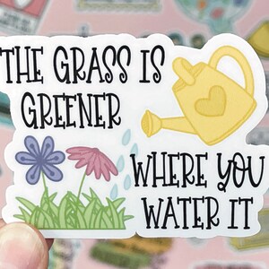 May include: A die-cut sticker featuring the black text THE GRASS IS GREENER WHERE YOU WATER IT. The design includes a yellow watering can with a heart, a purple flower, a pink flower, and green grass with small blue water droplets.