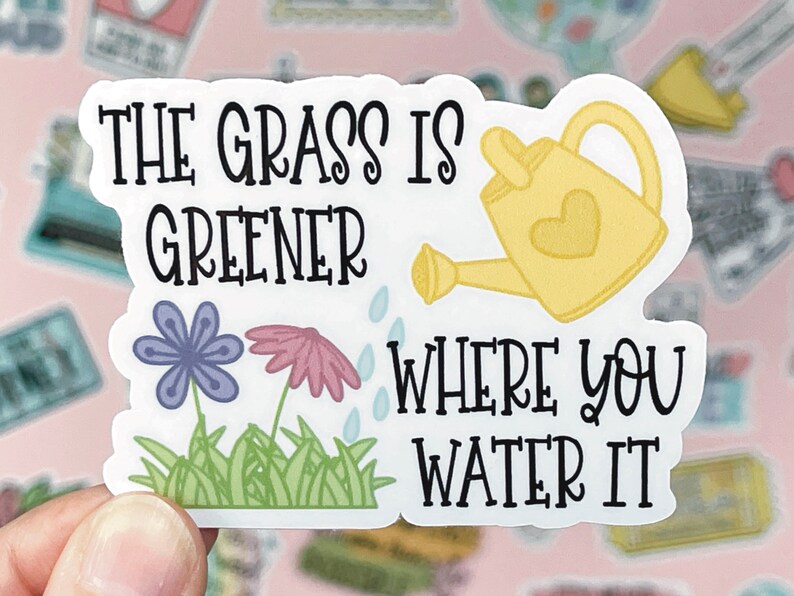 May include: A die-cut sticker featuring the black text THE GRASS IS GREENER WHERE YOU WATER IT. The design includes a yellow watering can with a heart, a purple flower, a pink flower, and green grass with small blue water droplets.