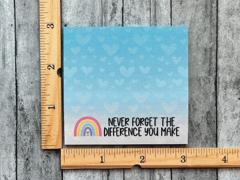 May include: A square note card with a light blue background featuring a pattern of faint hearts. The text reads "NEVER FORGET THE DIFFERENCE YOU MAKE" next to a small rainbow graphic. A wooden ruler shows the card is 3 inches wide and 3 inches tall.