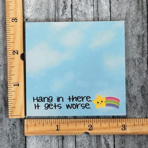 May include: A square light blue sticky note with a cloudy sky pattern, featuring the black text "Hang in there. It gets worse." and a small yellow star with a rainbow trail. A wooden ruler shows the note is approximately 3 inches wide and tall.