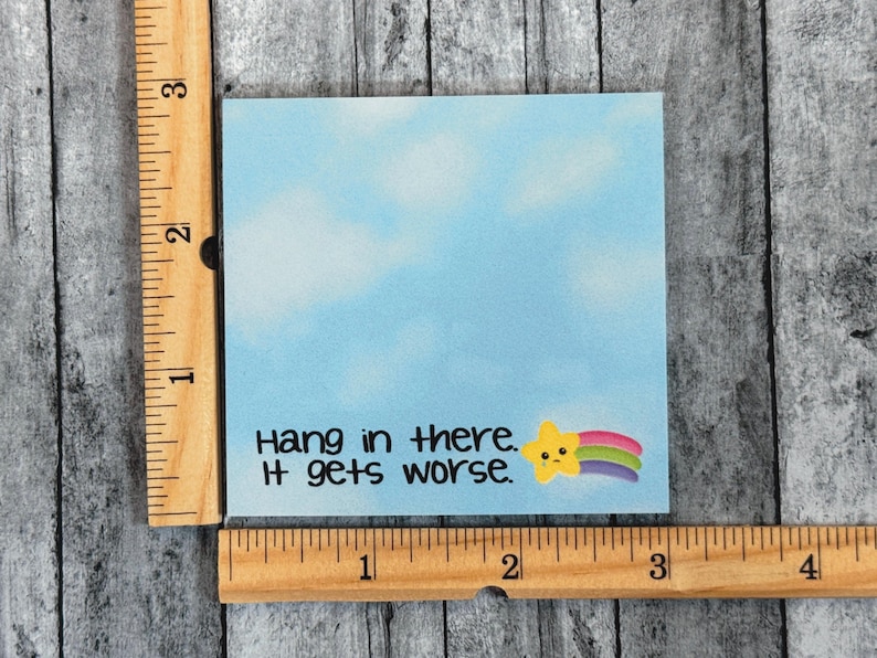 May include: A square light blue sticky note with a cloudy sky pattern, featuring the black text "Hang in there. It gets worse." and a small yellow star with a rainbow trail. A wooden ruler shows the note is approximately 3 inches wide and tall.