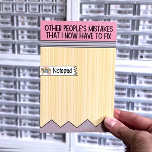 May include: A notepad with a pencil design. The top of the notepad is pink with the text "OTHER PEOPLE'S MISTAKES THAT I NOW HAVE TO FIX". The notepad has a light yellow wood grain design. The bottom has a gray zig-zag design.