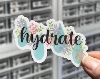 May include: A die-cut sticker featuring the word hydrate in black cursive script. The design includes light blue water drop shapes interspersed with small pink and purple floral illustrations and green leaves on a white background.