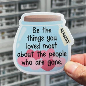 May include: A die-cut sticker shaped like a glass jar with a brown lid, filled with pink and red hearts. The jar features the black text: Be the things you loved most about the people who are gone. A small white tag labeled MEMORIES is attached.