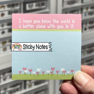 May include: A square sticky note with a light blue background and a green base featuring small pink and white heart-shaped flowers. The top section is pink with the white text: I hope you know the world is a better place with you in it.