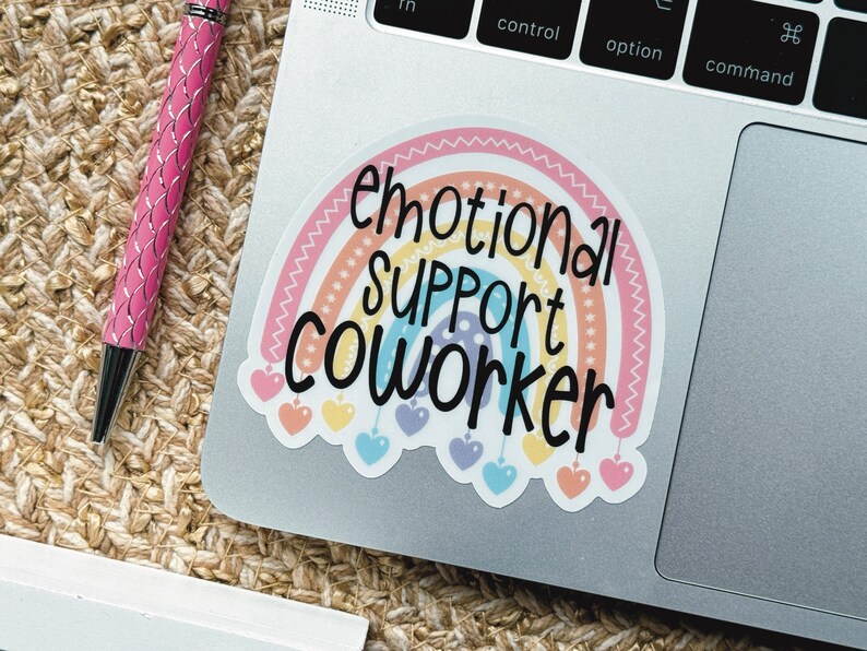 Puede incluir: Una pegatina de vinilo con un arco&iacute;ris de colores pastel con un borde festoneado y peque&ntilde;os colgantes en forma de coraz&oacute;n que cuelgan de la parte inferior. El texto "emotional SUPPORT COWORKER" est&aacute; impreso en letras negras sobre el arco&iacute;ris.