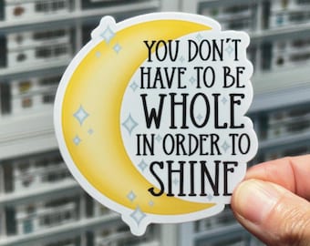 May include: A die-cut sticker featuring a yellow crescent moon shape with light blue four-pointed star accents. The black text reads, "YOU DON'T HAVE TO BE WHOLE IN ORDER TO SHINE," arranged in a stacked format across the center of the moon graphic.