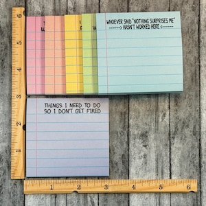 May include: A selection of colourful sticky note pads with lined pages. One pad has the text "THINGS I NEED TO DO SO I DON'T GET FIRED". Another pad has the text "WHOEVER SAID 'NOTHING SURPRISES ME' HASN'T WORKED HERE". A wooden ruler is next to the pads.