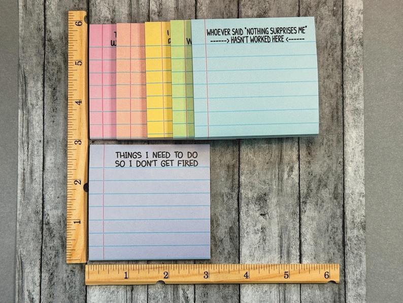 May include: A selection of colourful sticky note pads with lined pages. One pad has the text "THINGS I NEED TO DO SO I DON'T GET FIRED". Another pad has the text "WHOEVER SAID 'NOTHING SURPRISES ME' HASN'T WORKED HERE". A wooden ruler is next to the pads.